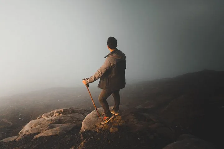 A hiker stands on a rocky mountain peak looking out at mist.