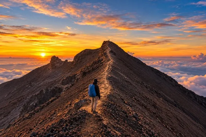 A hiker walks along a narrow rocky ridge towards the summit at sunrise.