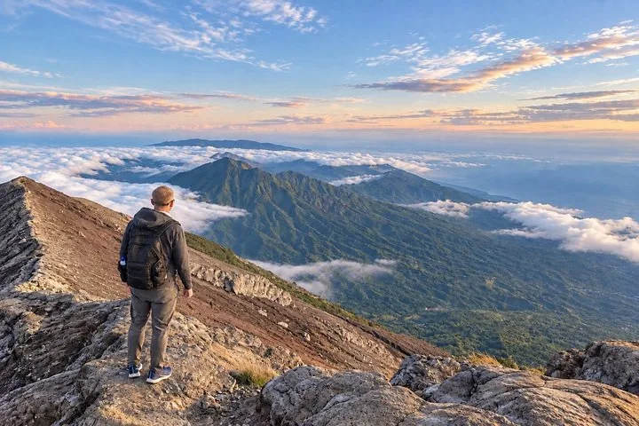 A hiker looks at a vast mountainous landscape above the clouds.