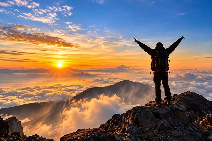 A hiker stands victorious on a rocky peak at sunrise above the clouds.