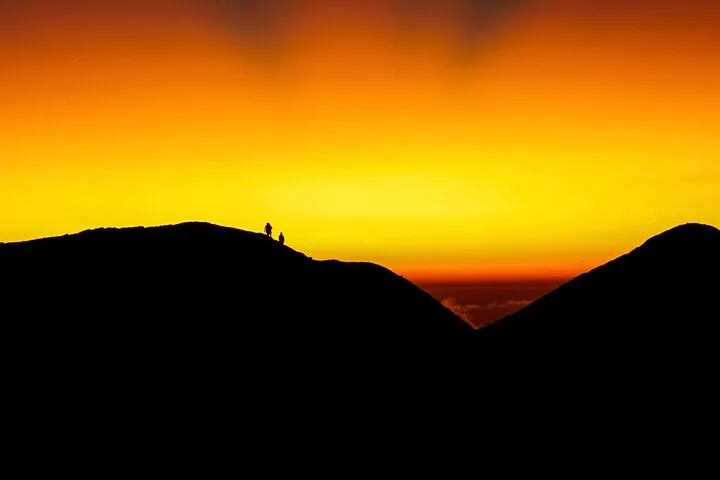 Silhouettes of two hikers on a mountain ridge against a bright orange sunrise.